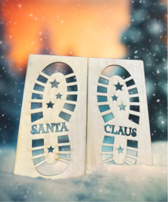 Two white wood stencils shaped like Santa's boots with the word 'SANTA' and stars printed on them, displayed against a snowy backdrop with a blurred Christmas light in the background.